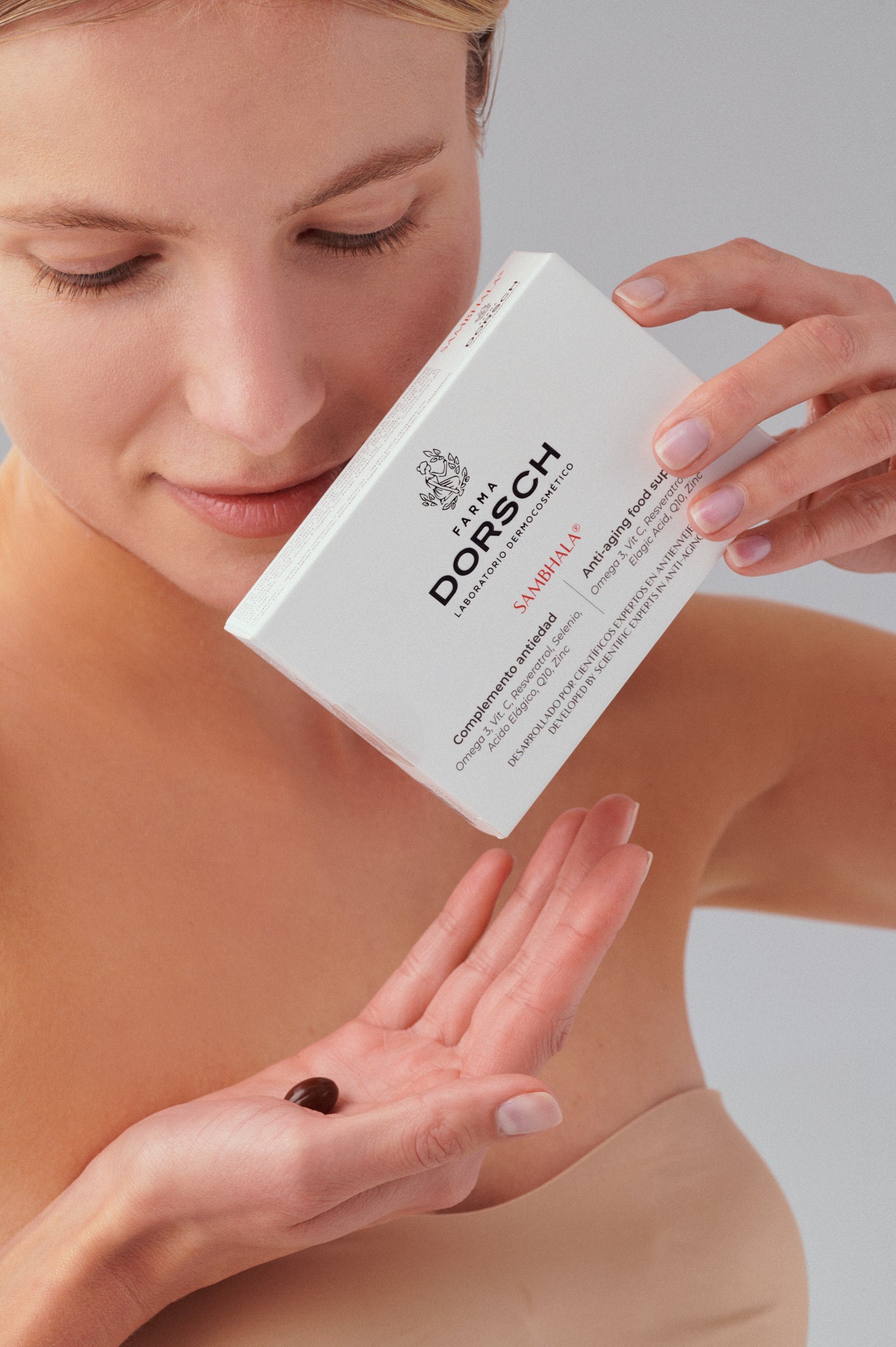 Mujer sosteniendo y observando una caja de SAMBHALA® FARMA DORSCH, complemento antiedad con Resveratrol, Coenzima Q10, Omega-3, Vitamina C, Zinc y Selenio, mientras con una cápsula en su mano.