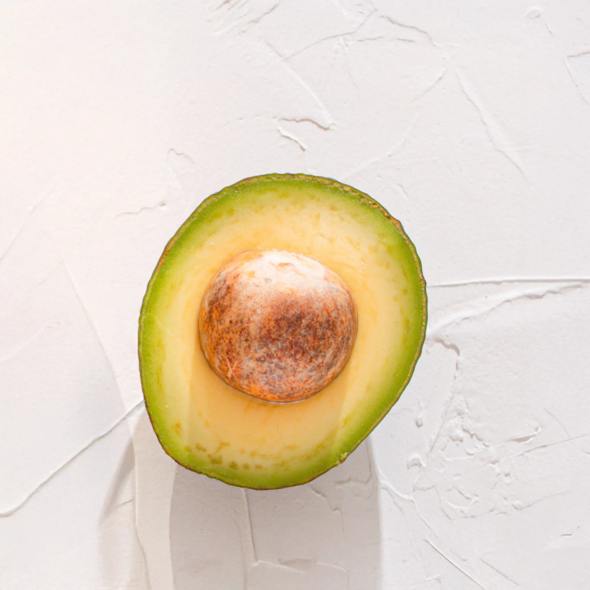Medio aguacate con hueso representando Ingrediente activo Vitamina E Farma Dorsch con propiedades hidratantes, antioxidantes y nutritivas para tratamiento facial y corporal sobre fondo blanco suave en cosmetica natural española