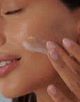 Mujer aplicando crema facial sobre la mejilla, primer plano que muestra el cuidado de la piel y textura del producto.