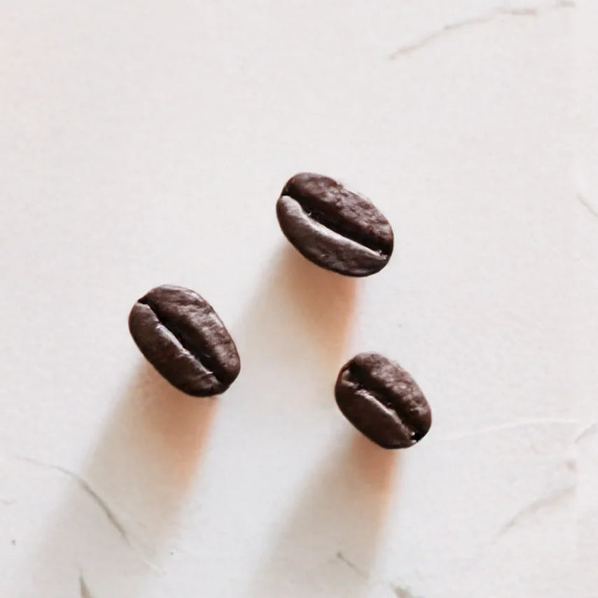 Tres granos de café representando Ingrediente activo Cafeína Farma Dorsch con propiedades estimulantes, antioxidantes y para mejorar la circulación en tratamiento facial y corporal sobre fondo blanco suave en cosmética natural española